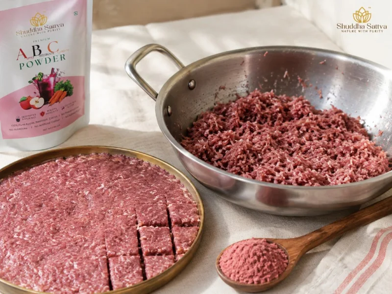 A kadhai of pink coconut burfi mix beside a brass thali of cut squares and a wooden spoon of ABC powder on cream linen